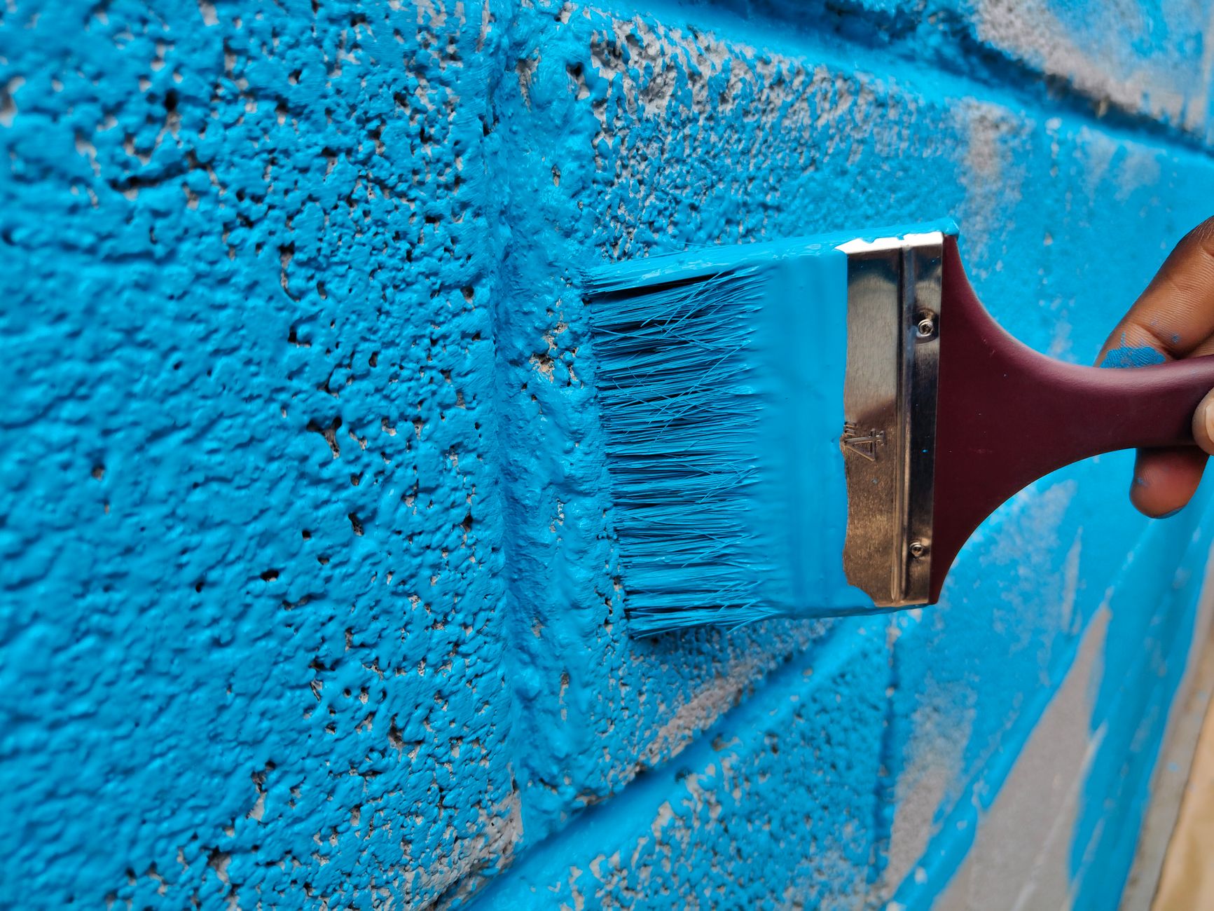A hand painting a brick wall with vibrant blue paint using a brush in Carlsbad, CA